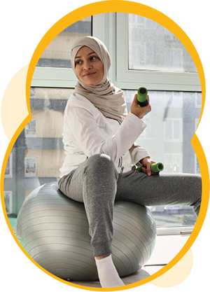 A person with medium skin, wearing a hijab, sits on a silver exercise ball. They are holding dumbbells in their hands.