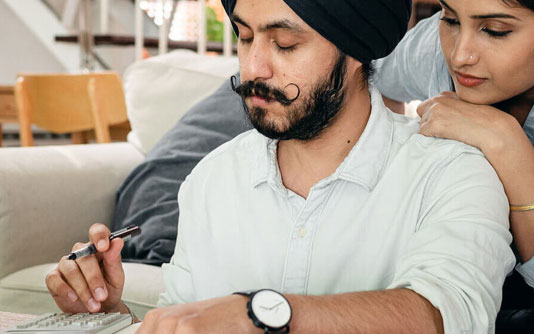 A man with a mustache wearing a turban holds a pen and calculator as a woman looks over his shoulder.