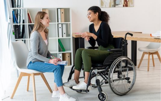 Positive young adult women talking – one seated on chair and one seated in wheelchair