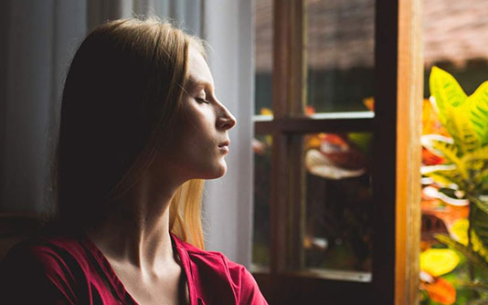 A woman eyes closed, in front of an open window looking calm and meditating