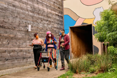 Three people with medium and medium-light skin wearing respirator masks and walking down a paved path together. One person is using a rollator for support and another is using a cane. Trois personnes &agrave; la peau moyennement claire ou l&eacute;g&egrave;rement claire, portant des masques respiratoires, marchent ensemble sur un chemin pav&eacute;. L&rsquo;une d&rsquo;elles s&rsquo;appuie sur un d&eacute;ambulateur et une autre sur une canne. 