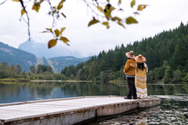 Two people wearing traditional Indigenous clothing standing on a dock, looking out at a lake, forest, and mountains in the distance. Deux personnes v&ecirc;tues de costumes traditionnels autochtones, debout sur un quai, contemplant un lac, une for&ecirc;t et des montagnes au loin. 