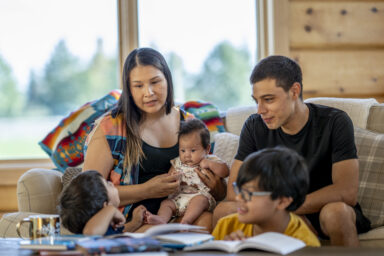 A family consisting of two parents, two school-age children, and a baby, all with medium-light skin, sitting on a couch smiling and having a conversation. Une famille de deux parents, deux enfants d&rsquo;&acirc;ge scolaire et d&rsquo;un b&eacute;b&eacute;, tous &agrave; la peau moyennement claire, assis sur un divan, souriants et en pleine conversation. 