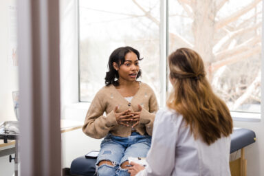 A patient with dark skin sitting on a medical examination table, having a conversation with a doctor with light skin. Un patient &agrave; la peau fonc&eacute;e, assis sur une table d&rsquo;examen m&eacute;dical, en train de discuter avec un m&eacute;decin &agrave; la peau claire.