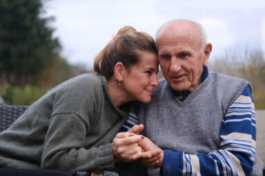 A young adult and an older adult, both with light skin, sitting on a bench together, holding hands. Un jeune adulte et une personne &acirc;g&eacute;e, tous deux &agrave; la peau claire, assis ensemble sur un banc, se tenant la main.