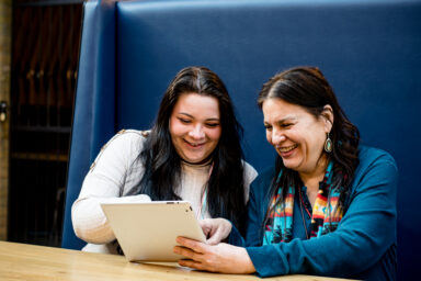 Two people with light skin sitting at a table, smiling and laughing together. One of them is holding a tablet device. Deux personnes &agrave; la peau claire assises &agrave; une table; elles sourient et rient ensemble. L&rsquo;une d&rsquo;elles tient une tablette. 