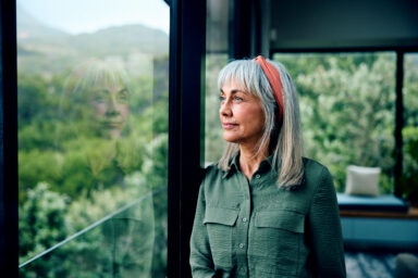 A person standing near a large window, looking out at the trees surrounding the house. Une personne debout pr&egrave;s d&rsquo;une grande fen&ecirc;tre, regardant les arbres qui entourent la maison. 